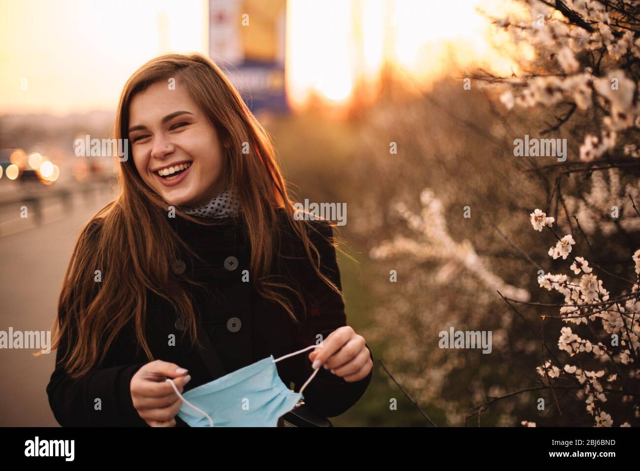 Happy cheerful young woman removing face medical mask while standing on street in city during sunset in spring Stock Photo