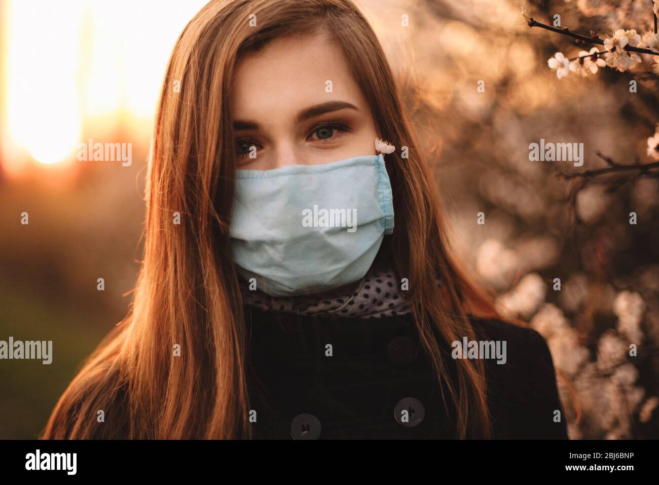 Portrait of cute happy teenage girl wearing face medical mask standing by flowering tree in park at sunset during spring Stock Photo