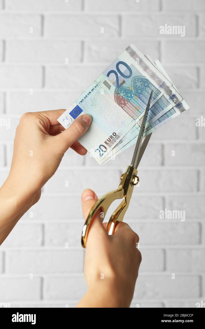 Hands with scissors cutting money on white brick wall background Stock