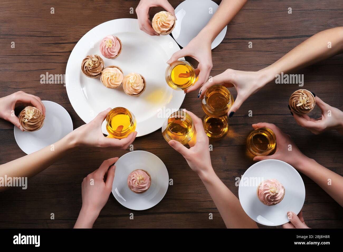 Female hands with glasses of tea and creamy cakes at wooden table, top view Stock Photo