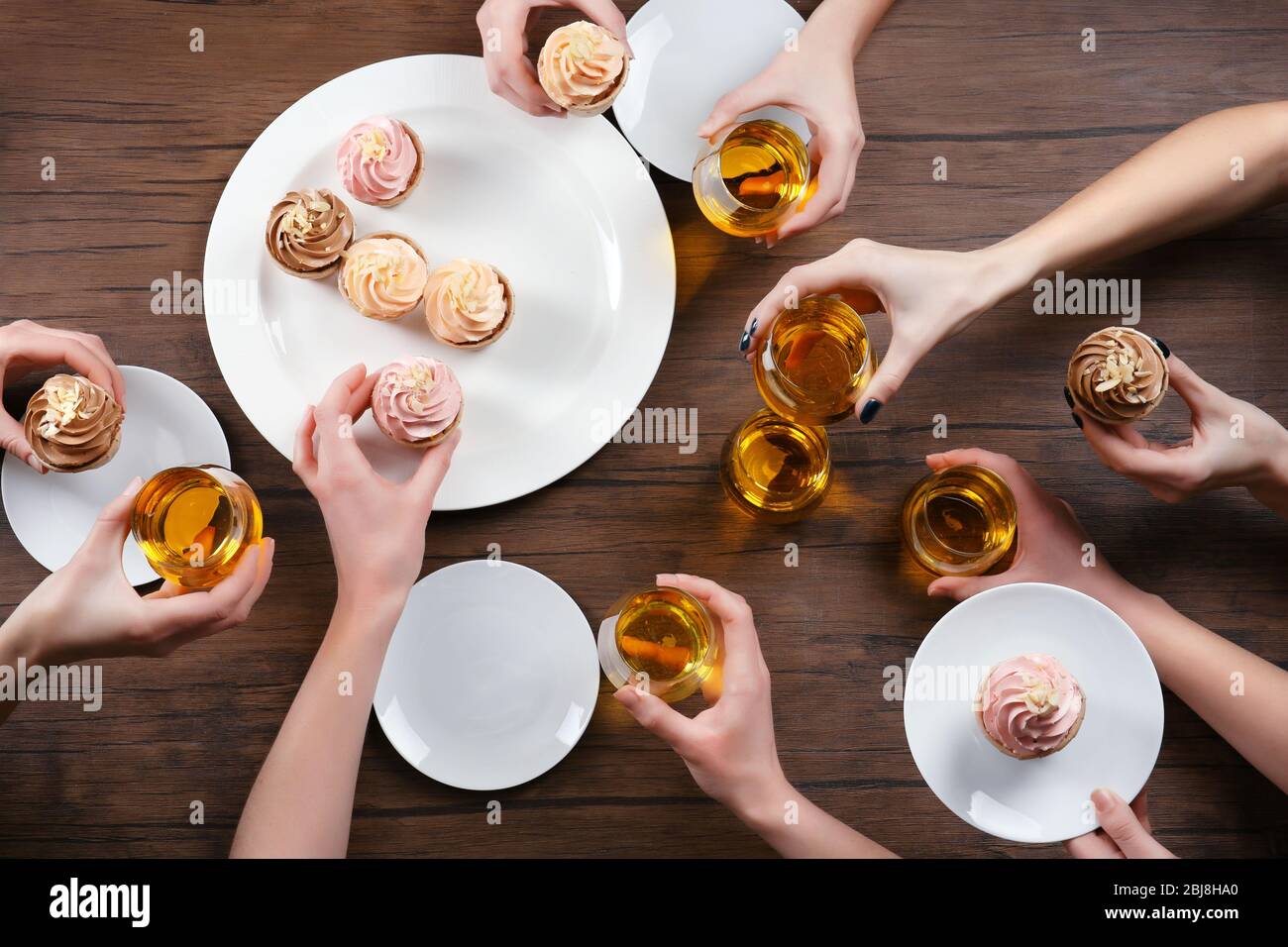 Female hands with glasses of tea and creamy cakes at wooden table, top view Stock Photo