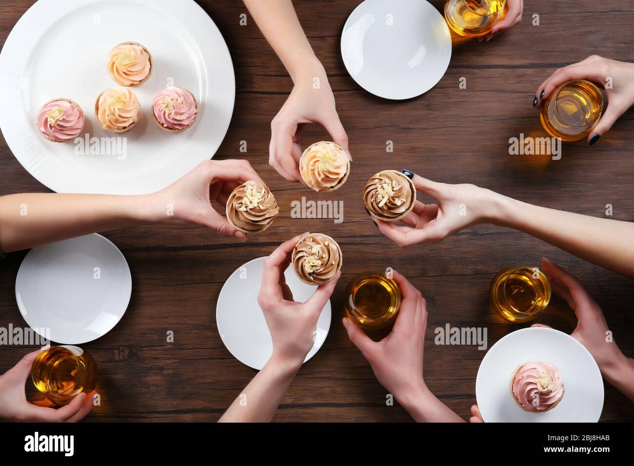 Female hands with glasses of tea and creamy cakes at wooden table, top view Stock Photo