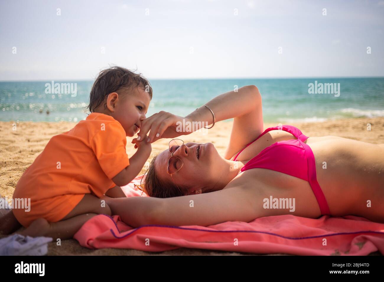 Little cute toddler is playing and pestering a young Caucasian mom who is lying on the sand of a beach. warm sea on a sunny day in the background. Stock Photo