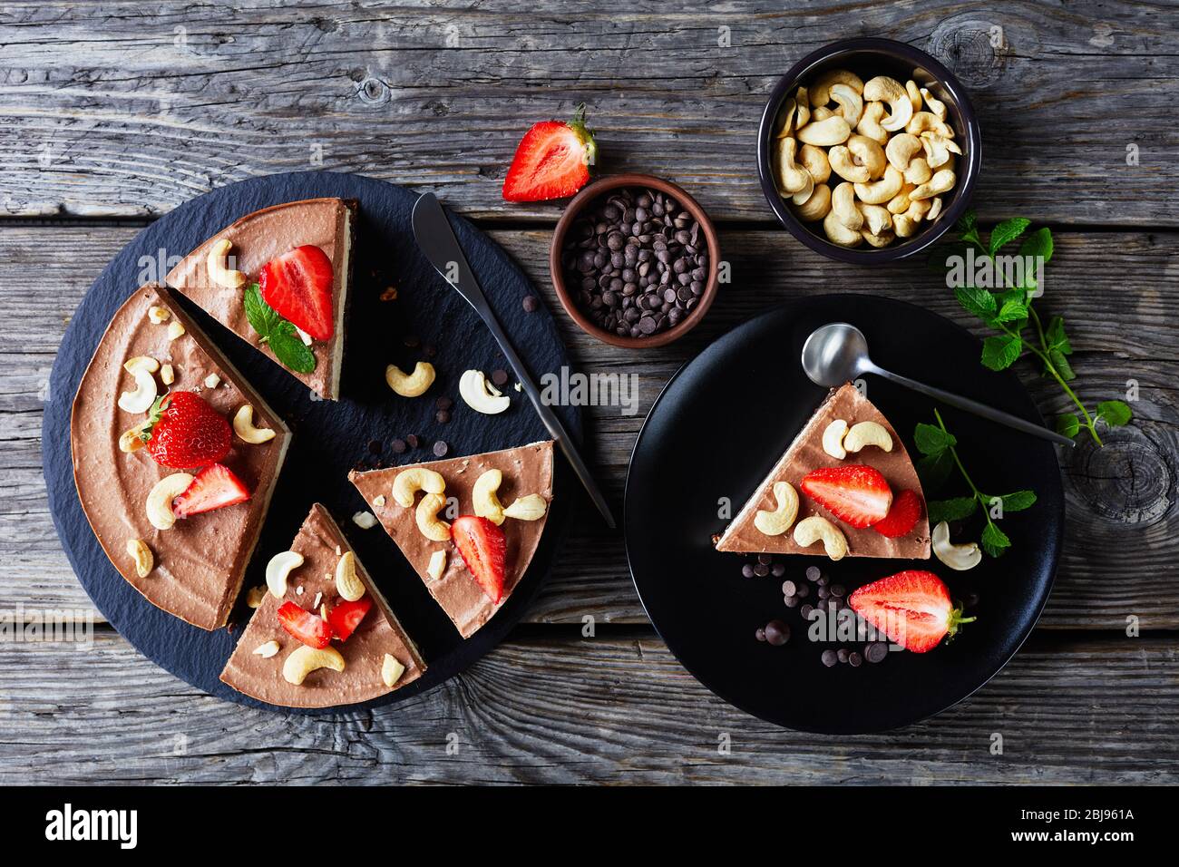 No-bake layered triple chocolate tart made of coconut cream and soaked cashews with the crust of raw dates, cocoa, and nuts with strawberries and mint Stock Photo