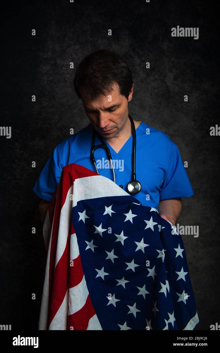 Sad/pensive male doctor in blue hospital scrubs with stethoscope, nursing the Stars & Stripes American flag close to his chest. Stock Photo