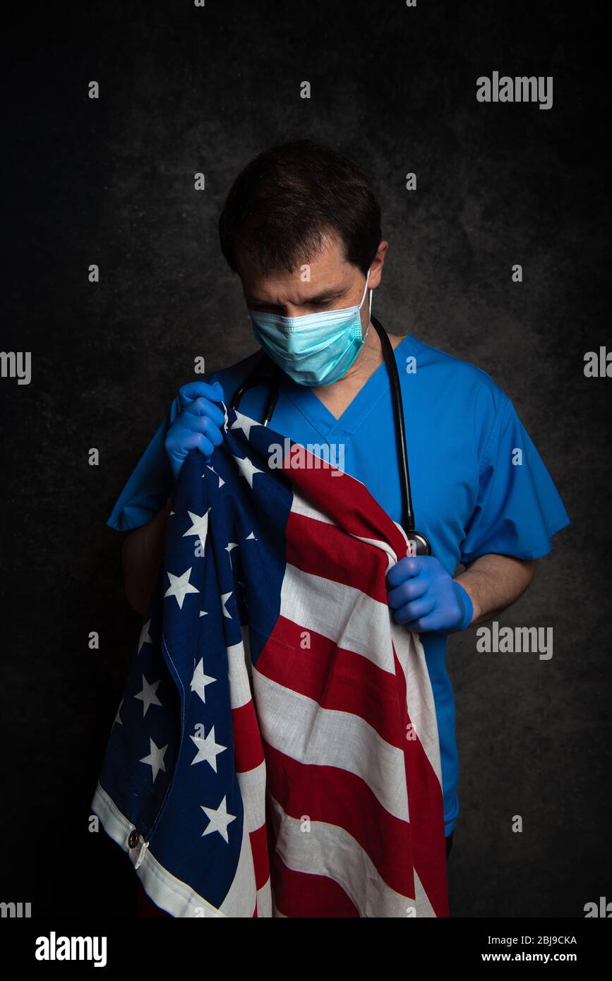 Sad/pensive, male doctor in blue hospital scrubs with face mask and stethoscope, nursing the Stars & Stripes American flag close to his chest. Stock Photo