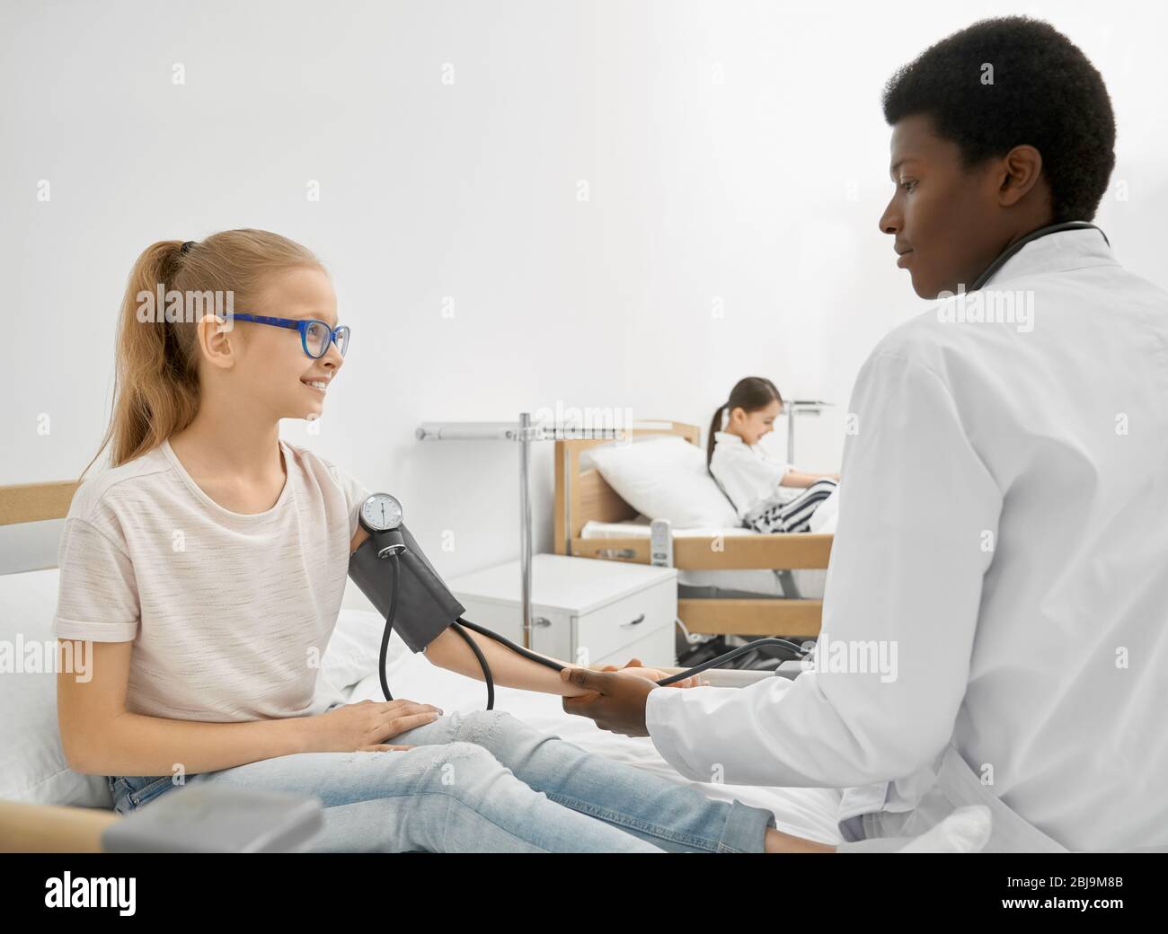 Side view of young african doctor measuring blood pressure of smiling schoolgirl in eyeglasses sitting on couch in hospital room. Little patient havin Stock Photo