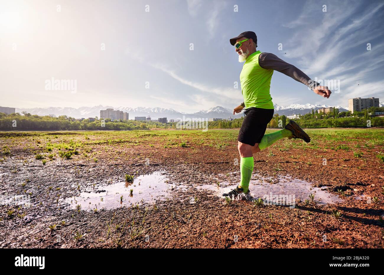 Man with grey beard running on the dirty trail with water splashes at mountain background in the morning. Healthy lifestyle concept Stock Photo