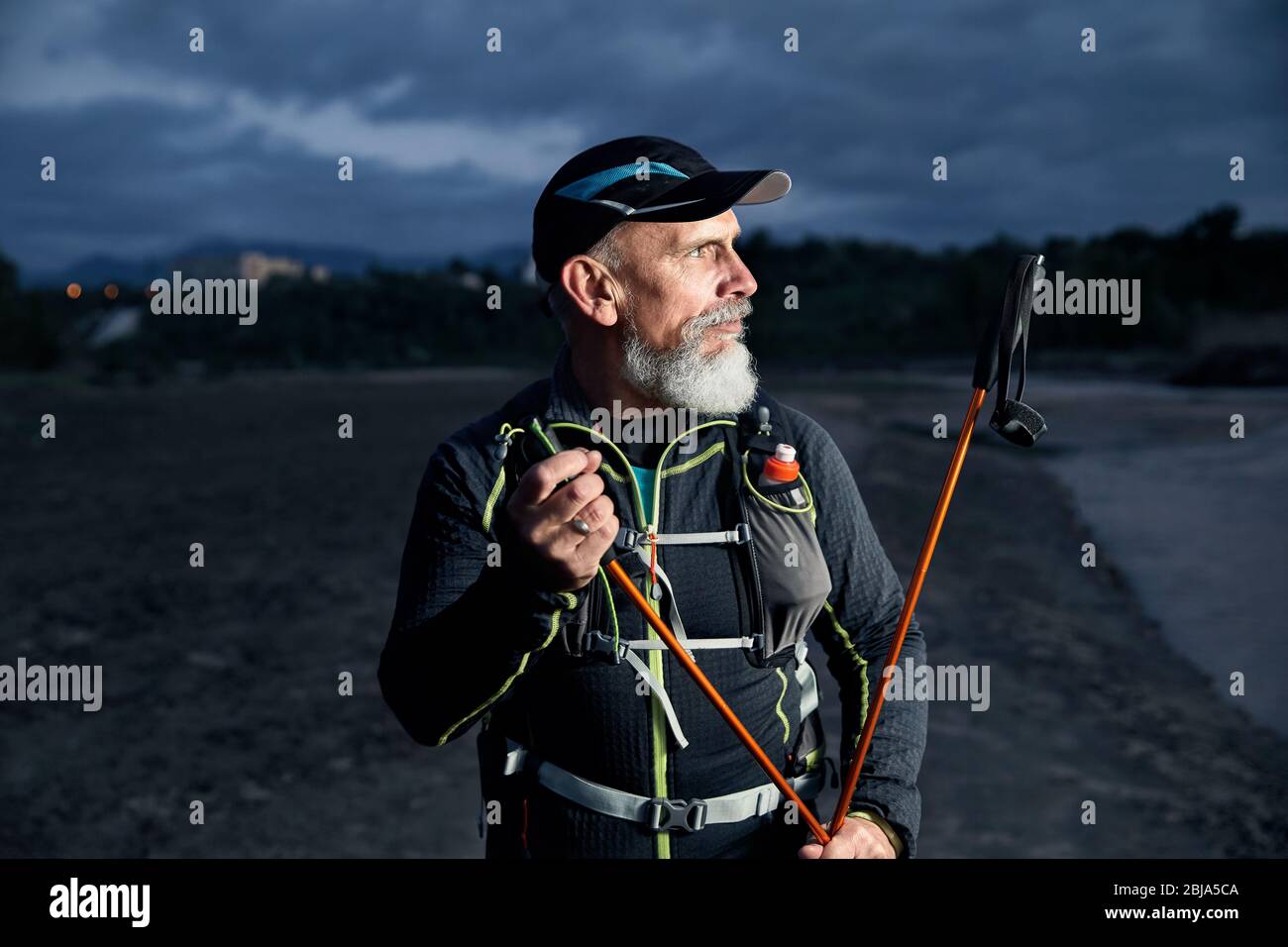 Portrait of elderly athlete man with grey beard and trekking poles at dark cloudy sky background Stock Photo