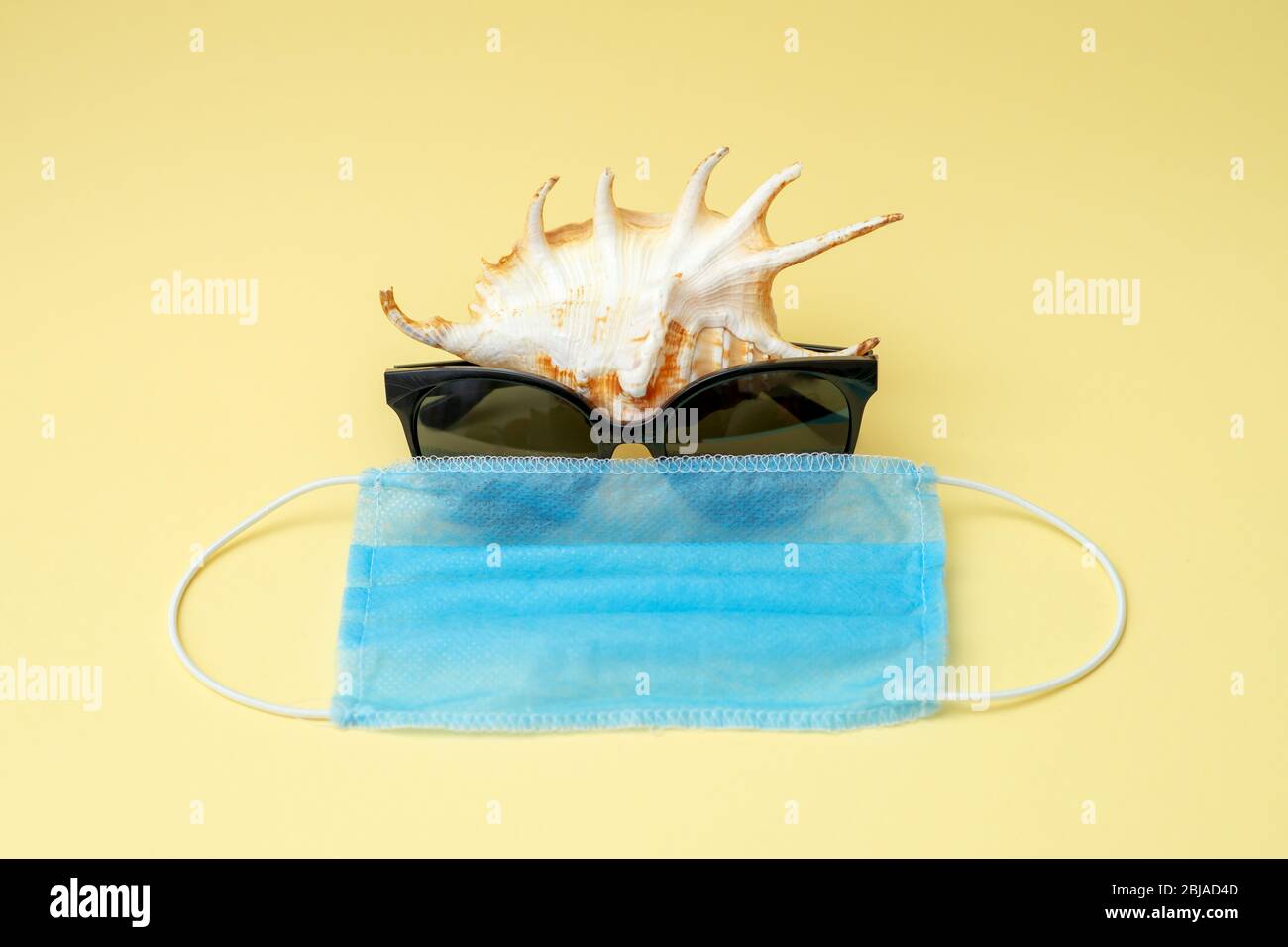 Seashell wearing sunglasses and medical mask on yellow background. Coronavirus or COVID-19 during traveling concept. Stock Photo