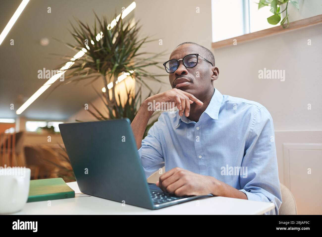Serious African businessman in eyeglasses sitting at the table in front of laptop and working online at office Stock Photo