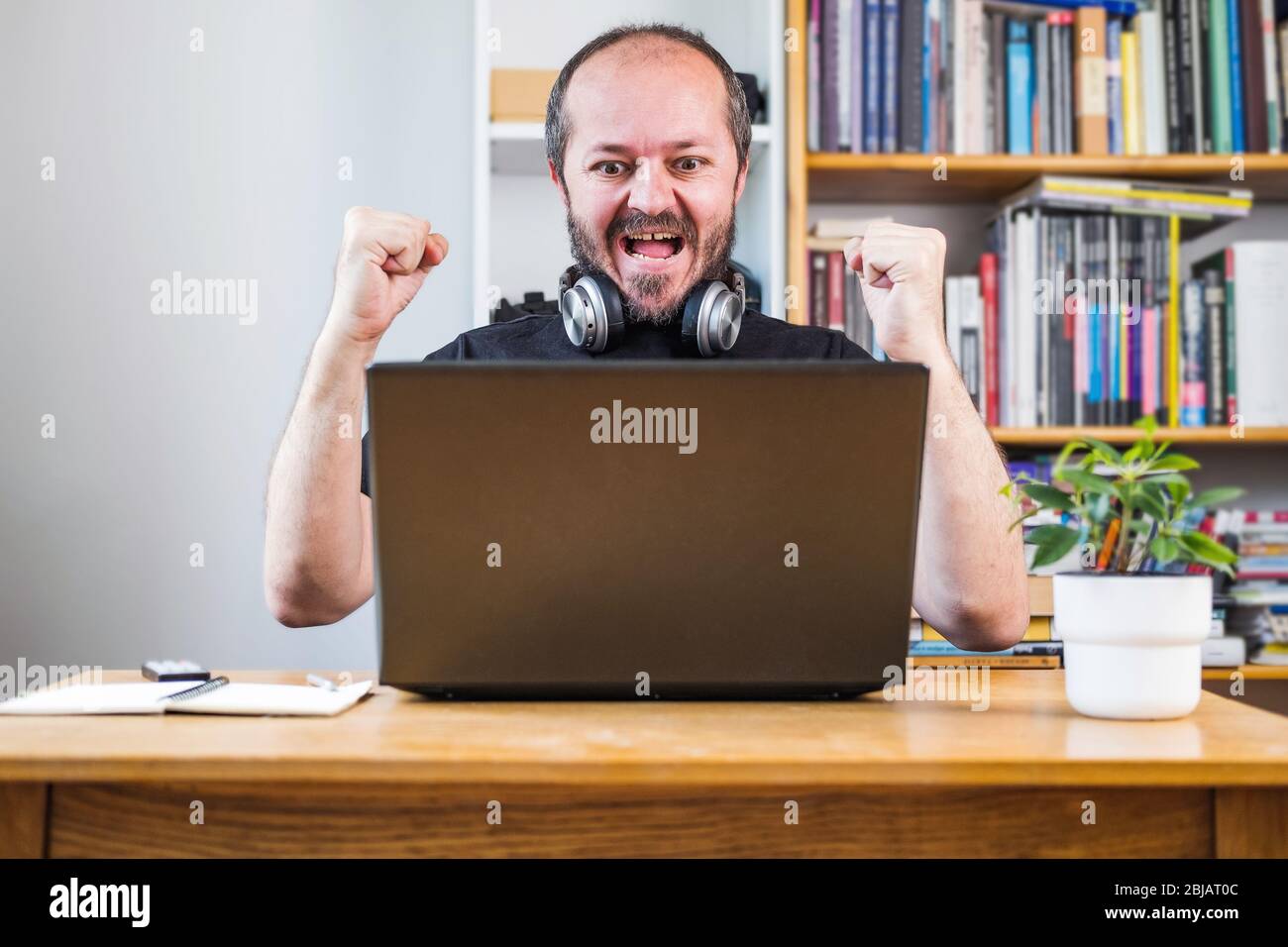 Man working from home office, happy and cheerful. Good news, new job. Bearded man on computer laptop behind vintage desk with flower in vase, with hea Stock Photo