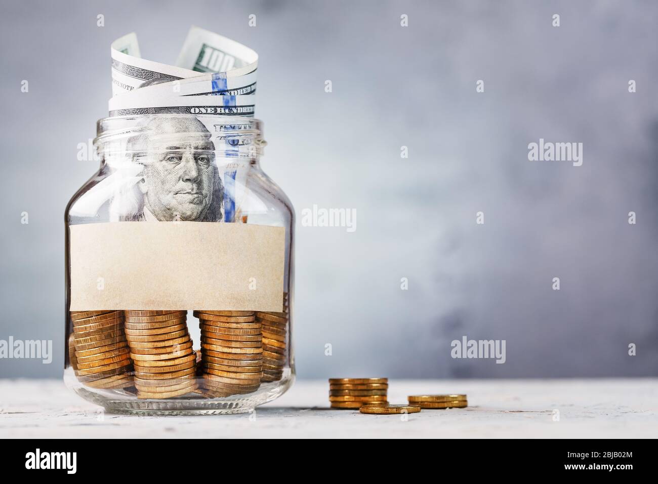 Glass Jar with gold coins, with a hundred-dollar bill and a sticker for text on a gray background with free space for text. Budget for investment in t Stock Photo