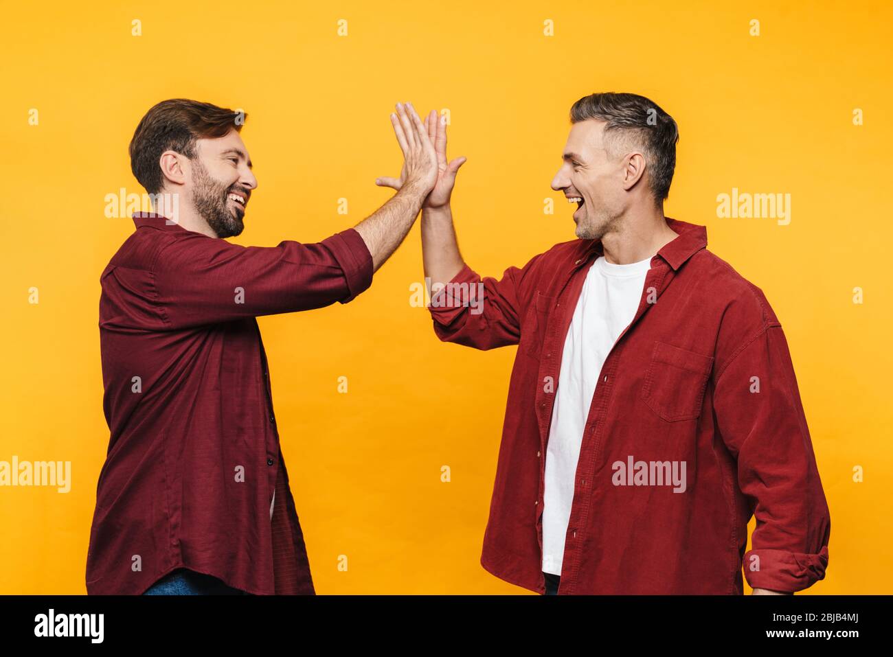 Image of two handsome men 30s in red shirts smiling and giving high five isolated over yellow background Stock Photo