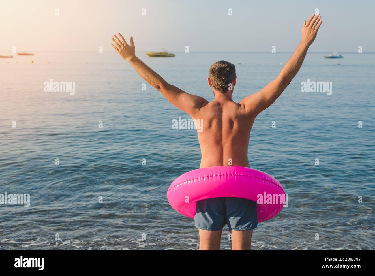 Happy man in pink with a big inflatable ring on the sea beach in summer sunny day. Back view. Stock Photo