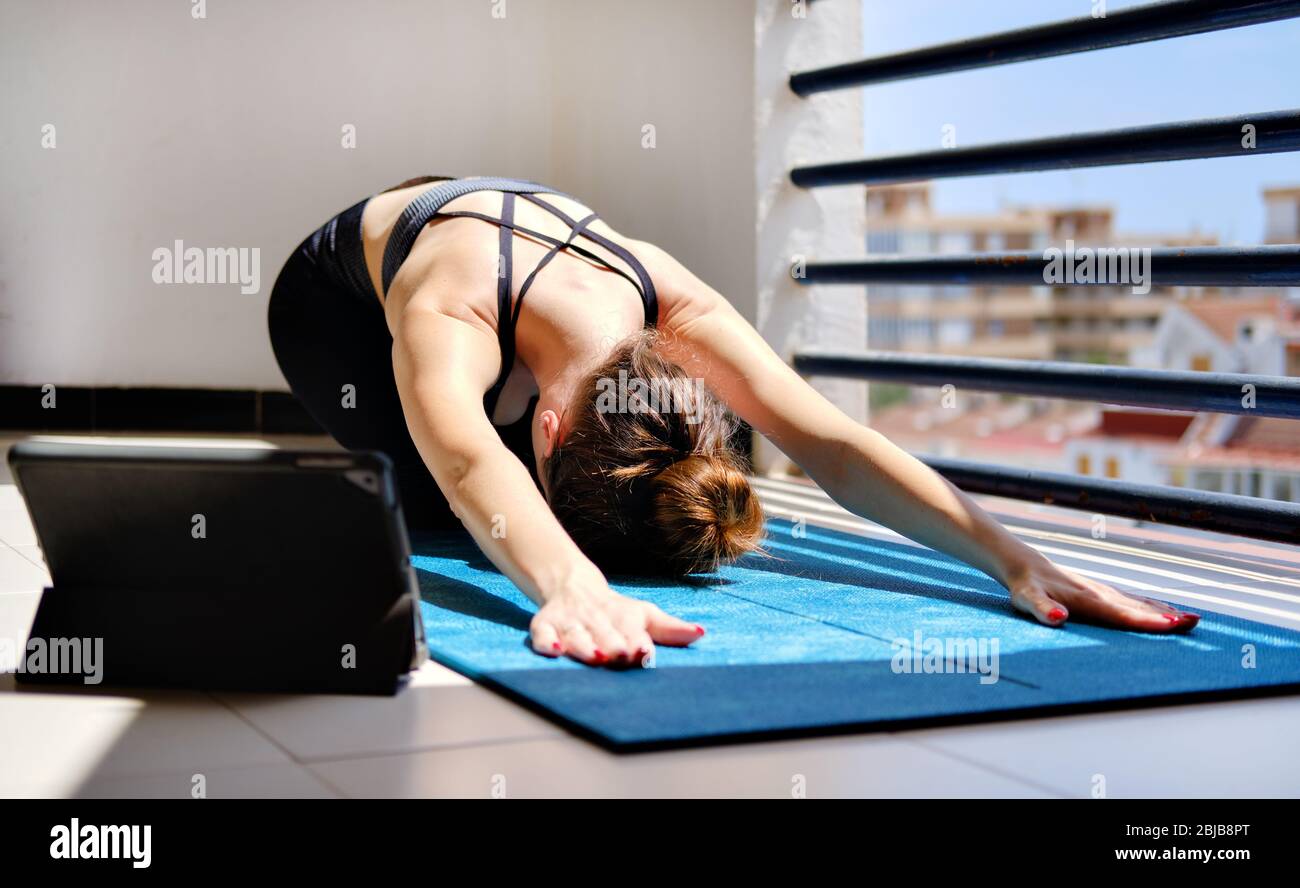 Woman perform yoga child pose on mat use on-line session on tablet. Workout at home in terrace due to self-isolation pandemic corona virus covid-19 Stock Photo