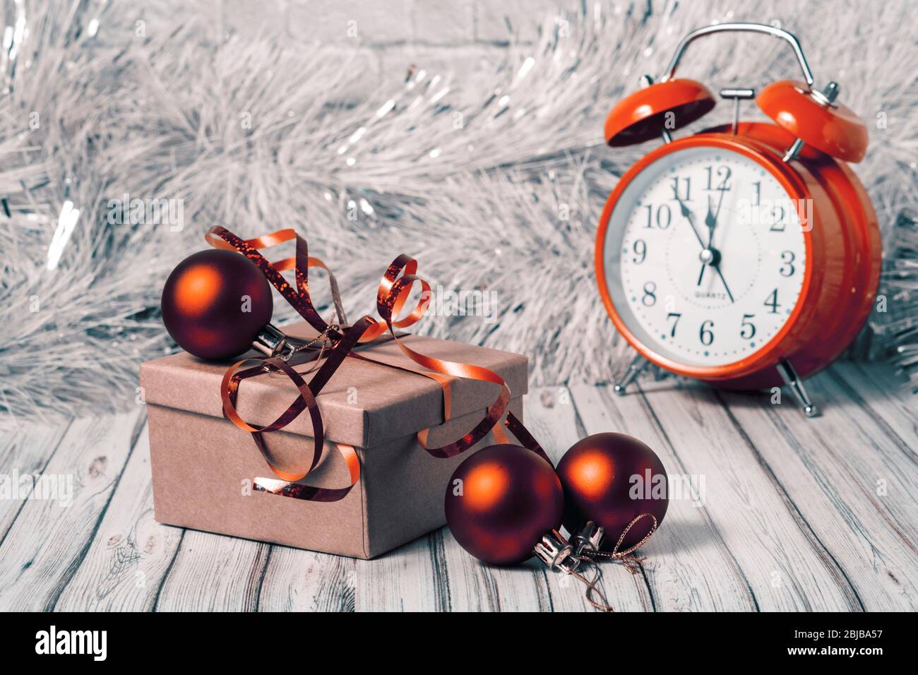 Gift box and blurred alarm clock on background on a wooden table decorated with a garland and red Christmas balls for the New Year or XMAS. Copy space Stock Photo