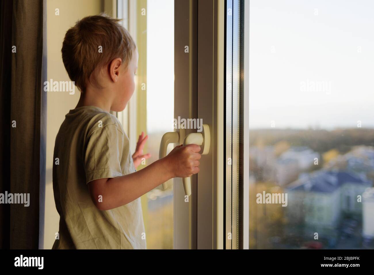 Boy looking through window to outside. Child opening window without parents. Dangerous situation Stock Photo