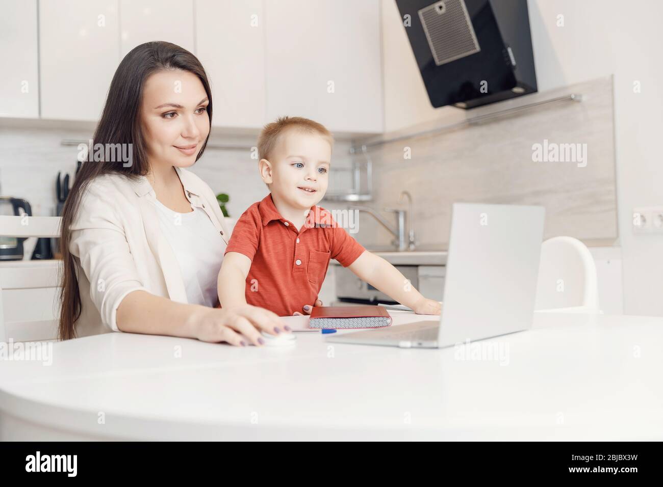 Woman on maternity leave working home online with laptop in kitchen with small child. Concept mom work while in quarantine isolation during Covid-19 Stock Photo