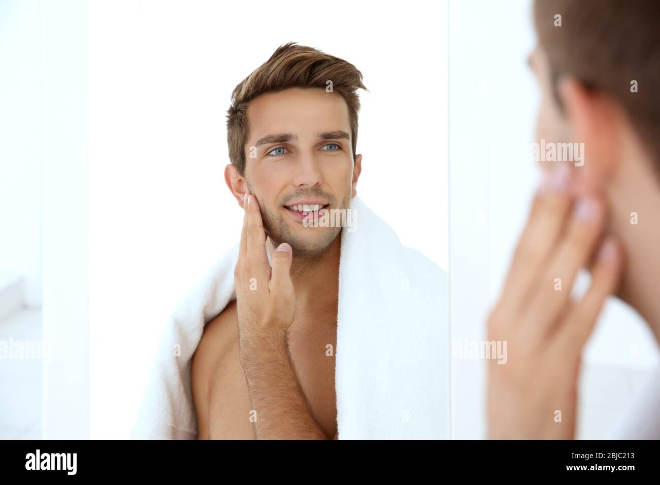 Young man standing in front of mirror and touching his face Stock Photo