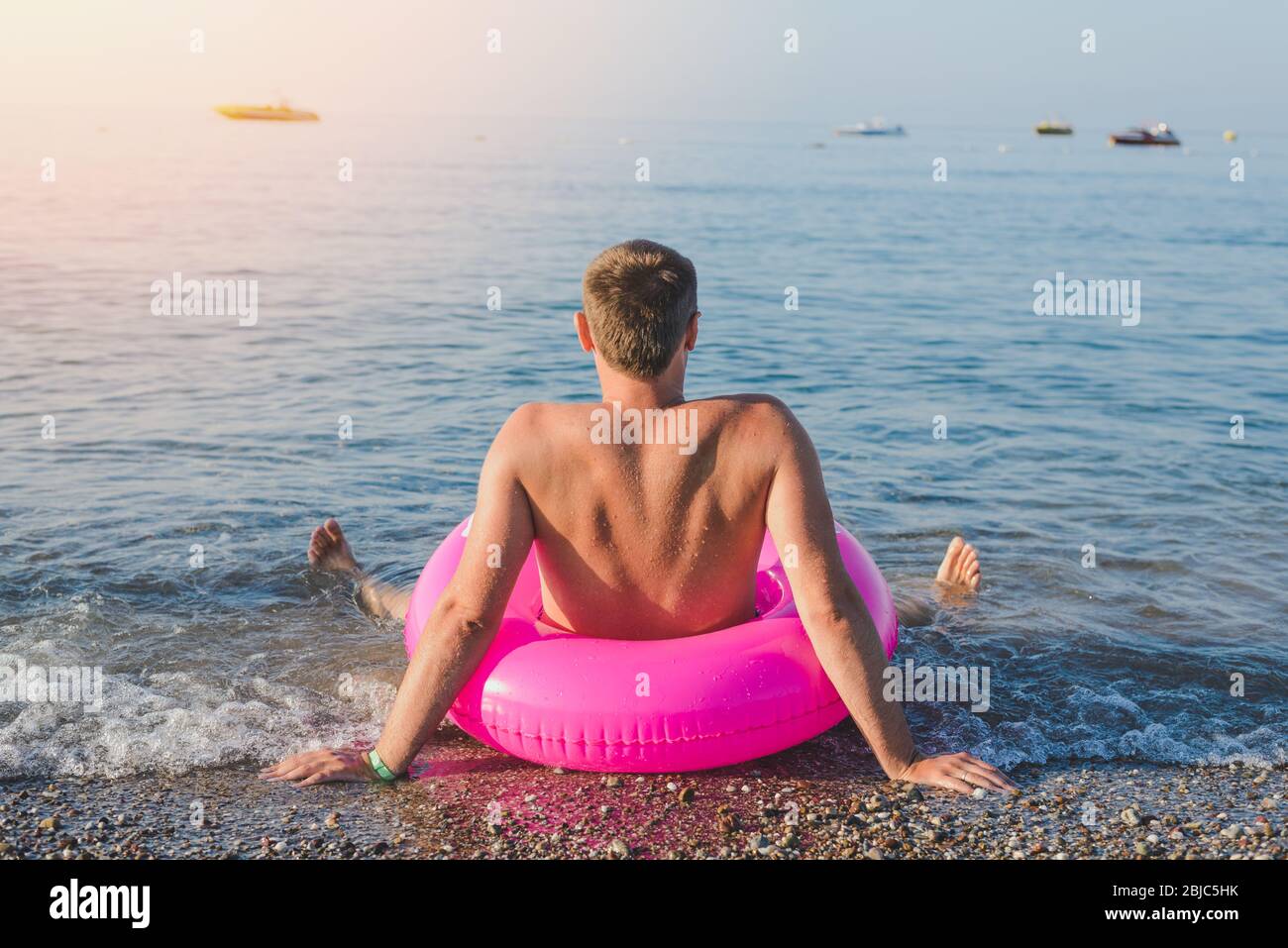 Happy man with big pink inflatable ring on the sea beach in summer sunny day. Back view. Stock Photo