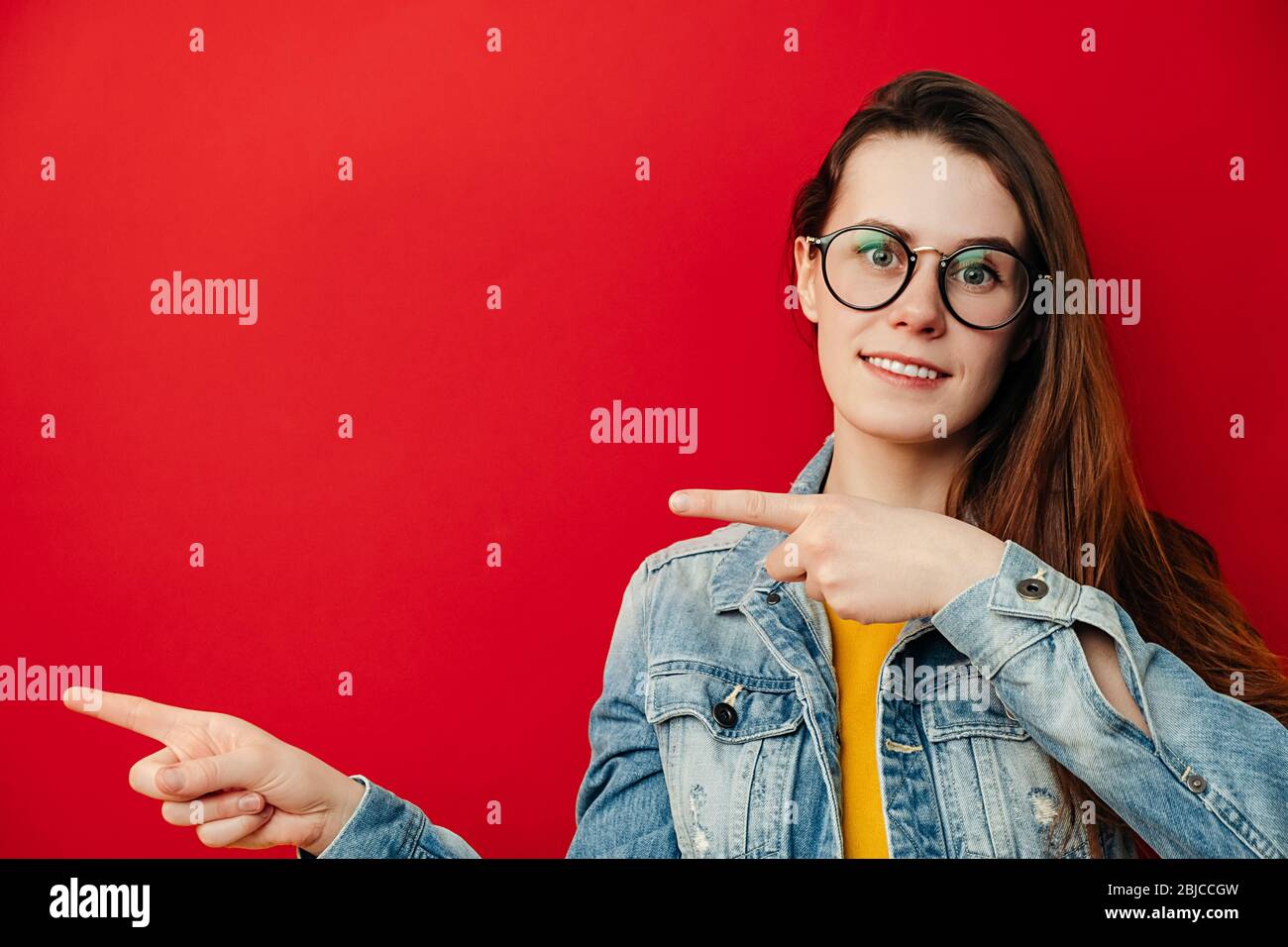 Beautiful brunette woman in eyeglasses, points with hands aside, shows copy space for your advertisement or promotional text, dressed in denim jacket, Stock Photo