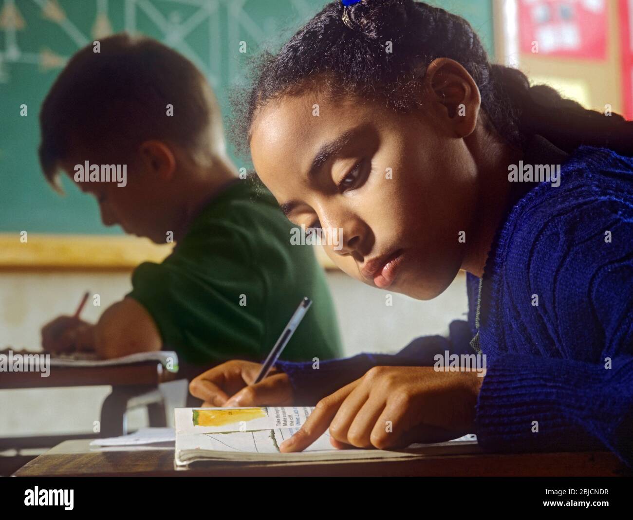 Junior girl school schoolgirl black African 9-11 years in classroom writing at her desk with blond boy & school geometry project blackboard behind Stock Photo