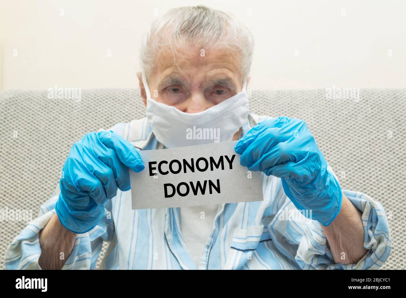 An elderly woman with a protective medical mask on her face and protective gloves hold paper with text ECONOMY DOWN. 2020 world economic crisis caused Stock Photo