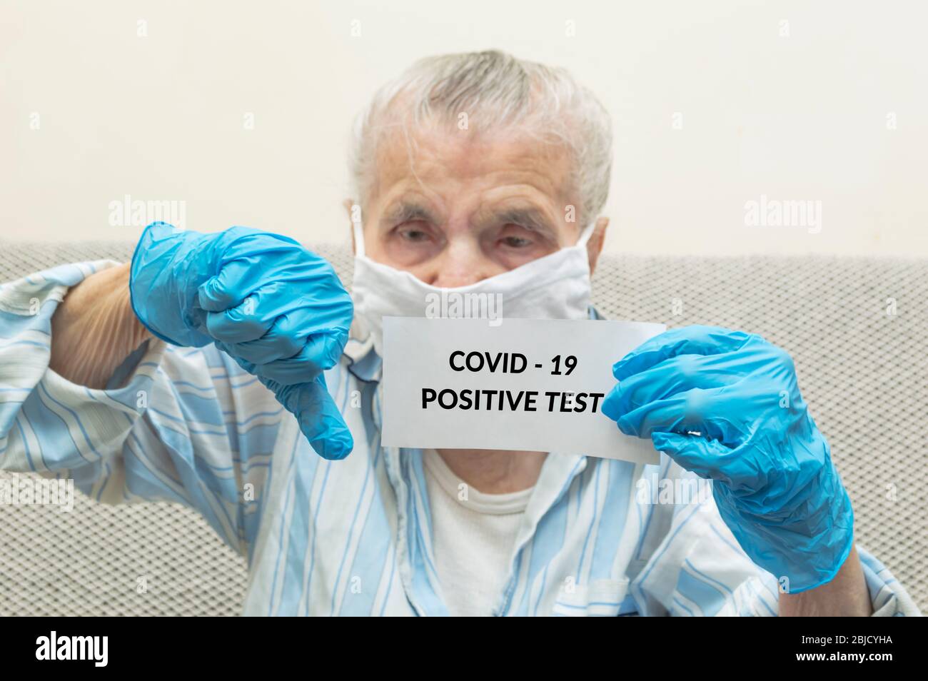 Elderly woman with protective mask holding a paper with a positive result on coronavirus. Sick granny looking into camera showing sad emotions. Concep Stock Photo
