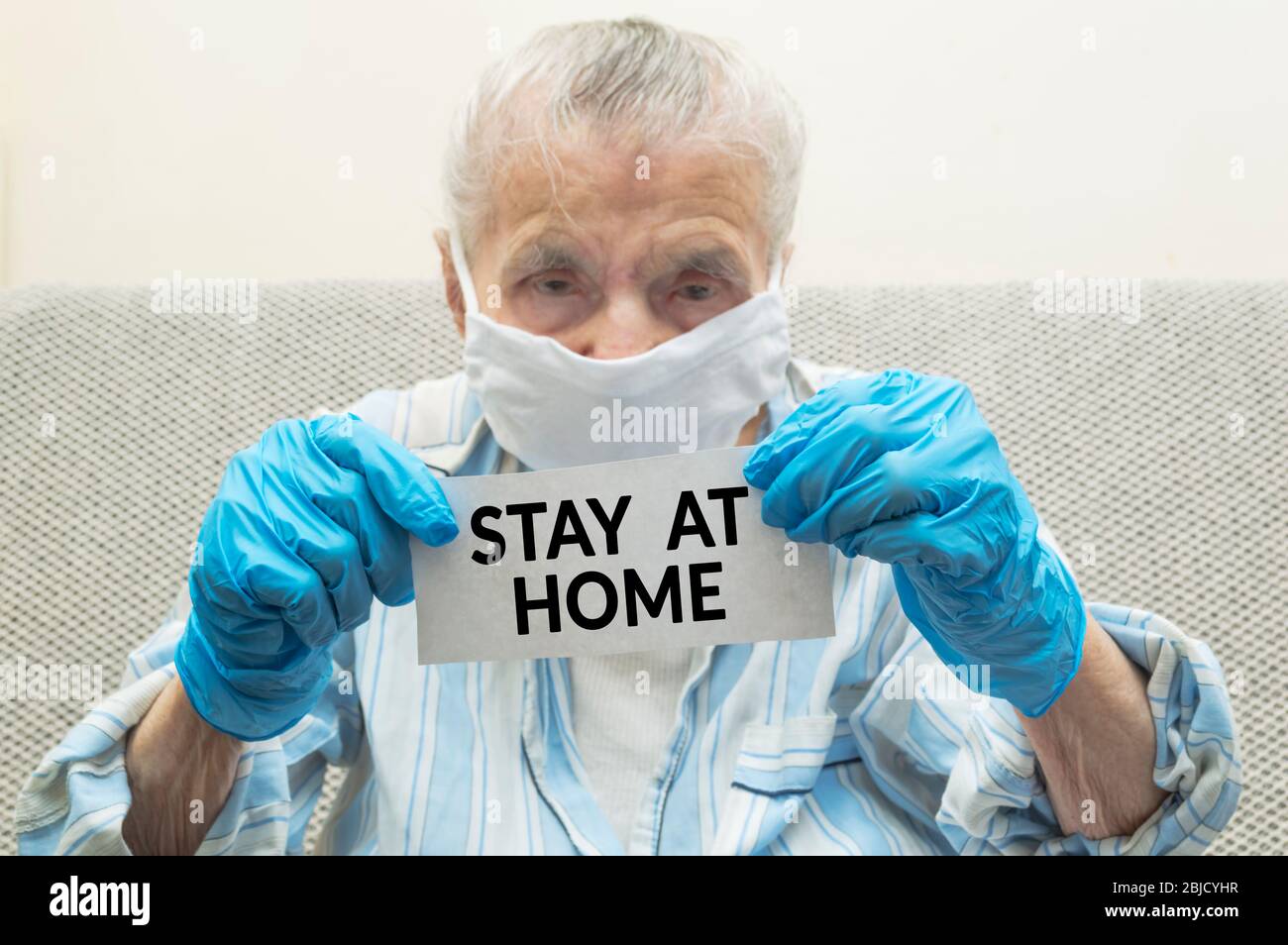 An elderly woman with a protective medical mask on her face and protective gloves hold paper with text the inscription STAY AT HOME. The concept - qua Stock Photo