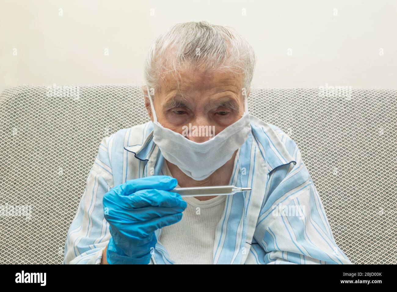 An elderly woman in a respiratory protection mask at home measures the temperature with a thermometer. Health of retirees. Isolation of the elderly, q Stock Photo
