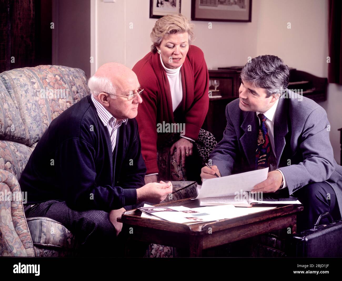 Mature retired elderly couple at home 1990s era, talking to salesman sales person man, with variety of paperwork on table. Financial planning, home improvements, legal services, will wills writing etc. Archive 1990's home services salesman Stock Photo