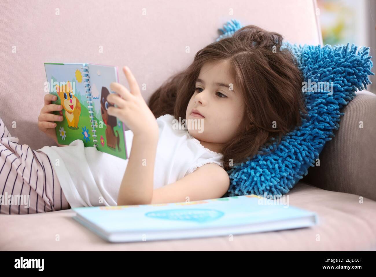 Funny little girl reading book while lying on sofa at home Stock Photo ...
