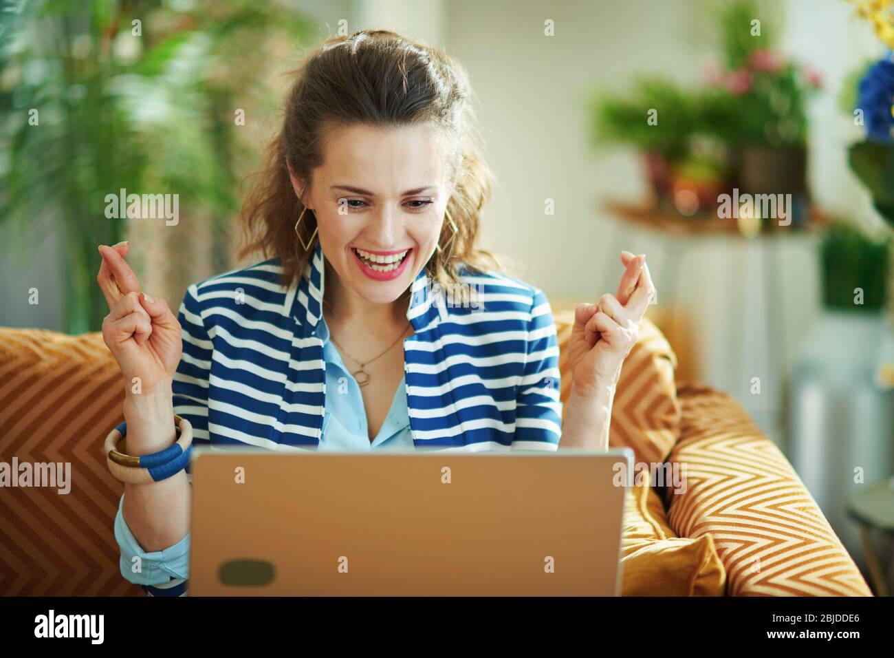 excited trendy housewife in blue blouse and striped jacket with crossed fingers sitting on couch and looking at laptop in the modern house in sunny da Stock Photo