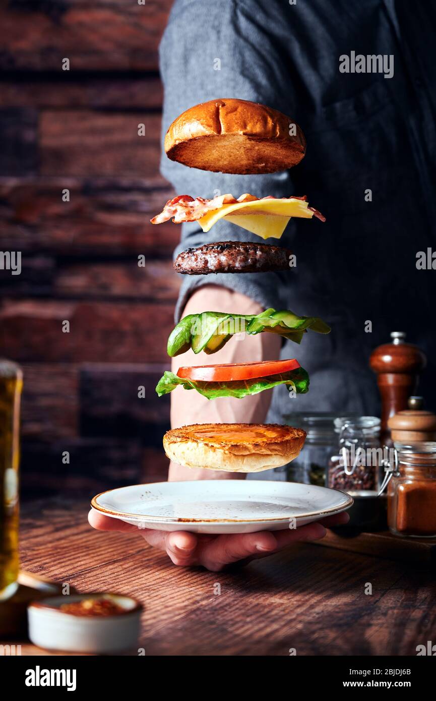 Flying beef burger with baby gem lettuce,tomatoes,dill pickles,beef patty,mature cheddar cheese bacon and sriracha mayo. wooden backdrop and counter Stock Photo