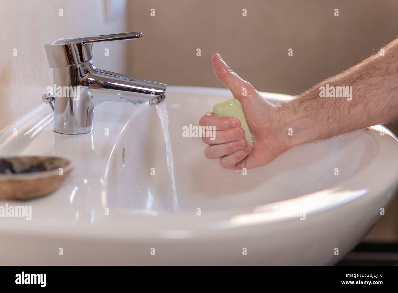 Hand of young caucasian man holding a bar of soap and showing thumb up gesture. Water tap is running. Personal hygiene, economical usage of water Stock Photo