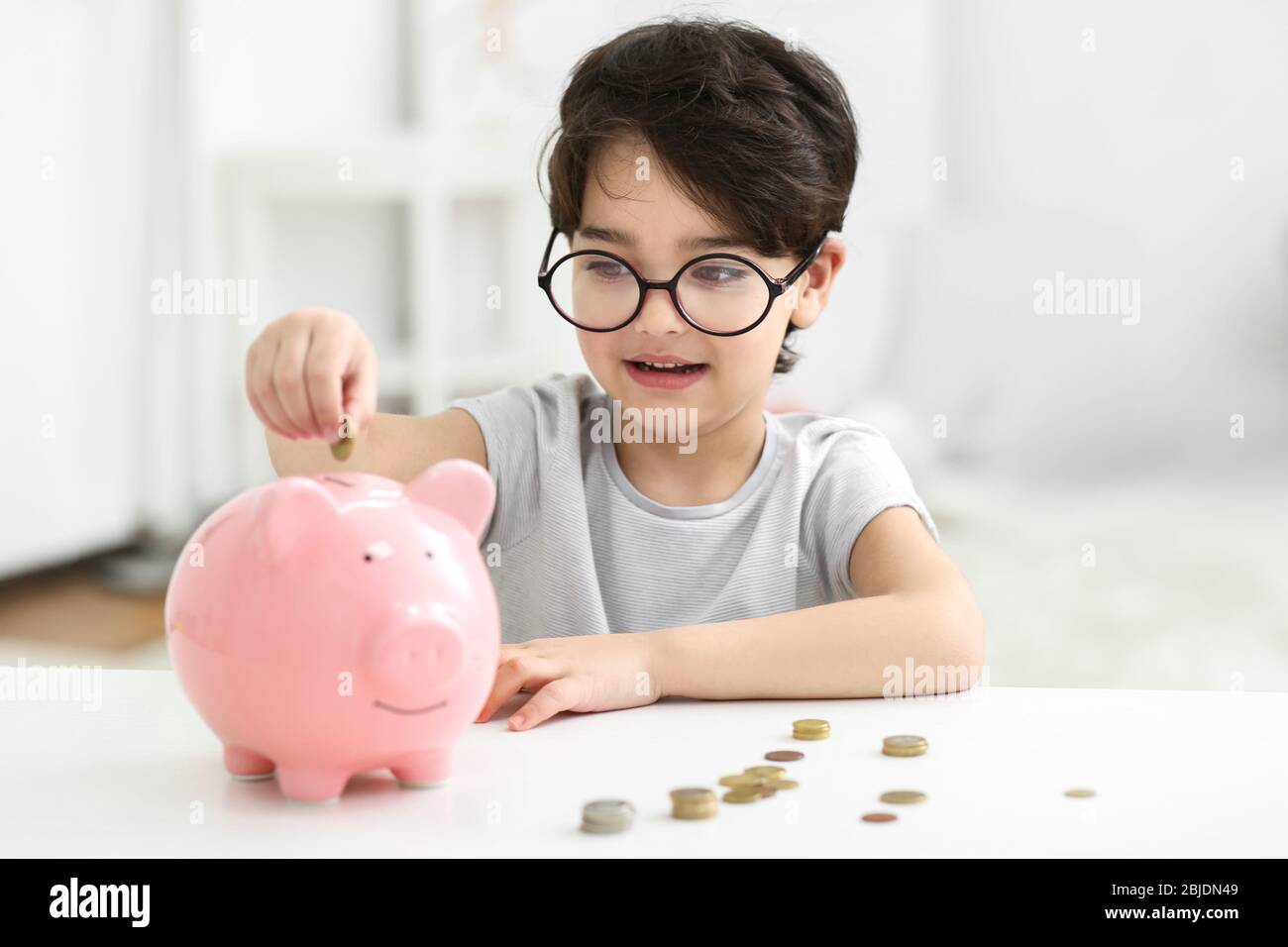 Portrait of cute little boy in glasses putting coin into a piggy bank ...