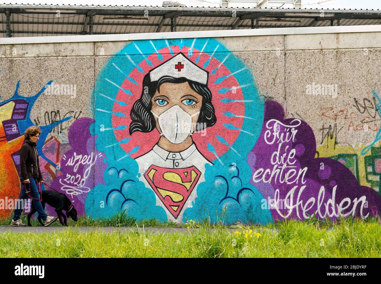 Hamm/Germany, April 29th, 2020: Poeple passing a coronavirus graffiti by street artist Kai „Uzey“ Wohlgemuth showing 'Super-Nurse' with a protection mask on a wall in Hamm, Germany. Scripture says: 'For the real heroes'    ---   Hamm, 29.4.2020 - Corona-Kunst. Das Graffiti „Super-Nurse“ an einer Wand in Hamm. Eine Krankenschwester mit Mundschutz und Supermann-Logo hat der Graffiti-Künstler Kai „Uzey“ Wohlgemuth aus Hamm den echten Helden in der Corona-Krise gewidmet. Stock Photo