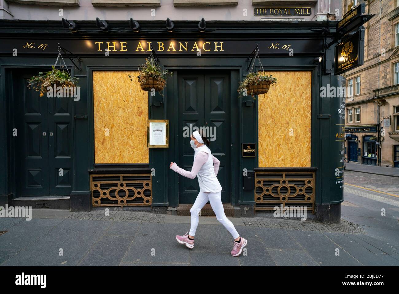 Edinburgh, Scotland, UK. 29 April 2020. Views of Edinburgh Old Town as coronavirus lockdown continues in Scotland. Streets remain deserted and shops and restaurants closed and many boarded up. Scottish Government now recommends public to wear face masks. Female jogger wearing face mask runs past a closed and boarded up Albanach pub on the Royal Mile. Iain Masterton/Alamy Live News Stock Photo