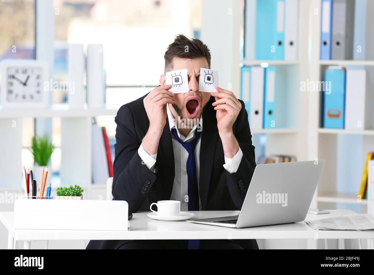 Young business man with fake eyes painted on paper stickers sleeping at  workplace in office Stock Photo
