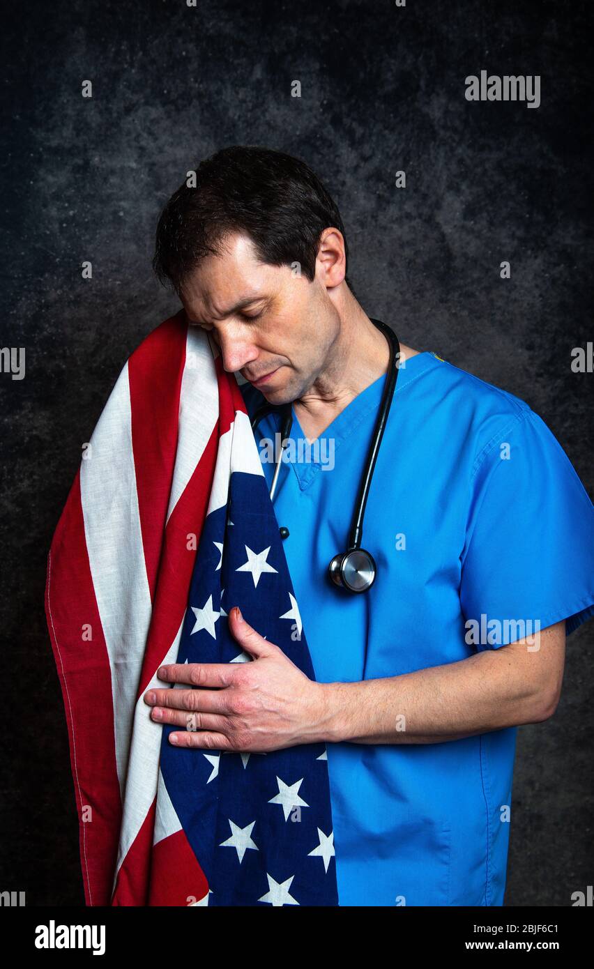 Sad/pensive male doctor in blue hospital scrubs with stethoscope, nursing the Stars & Stripes American flag close to his chest, against a dark studio. Stock Photo