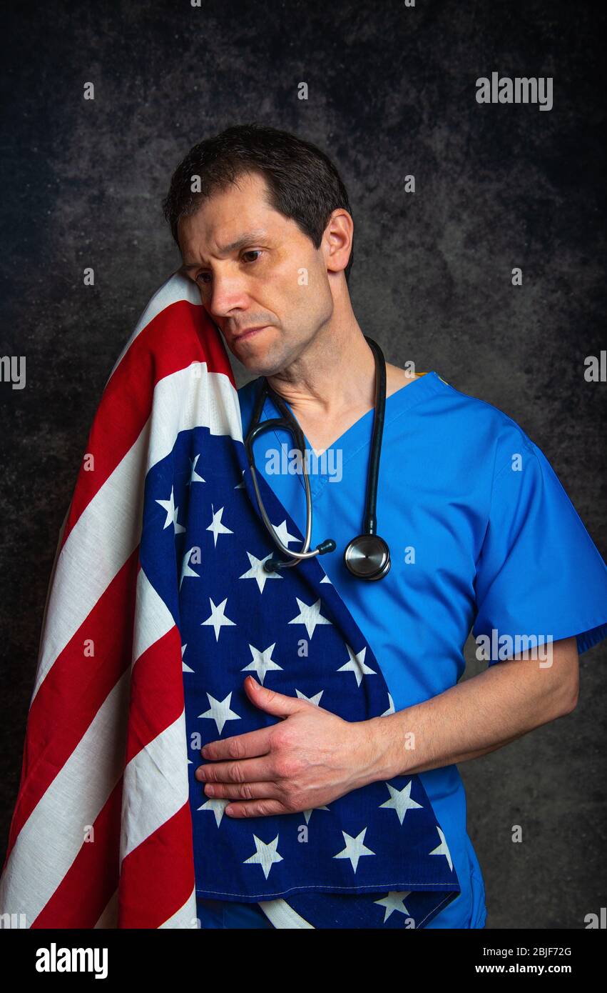 Sad/pensive male doctor in blue hospital scrubs with stethoscope, nursing the Stars & Stripes American flag close to his chest, against a dark studio. Stock Photo