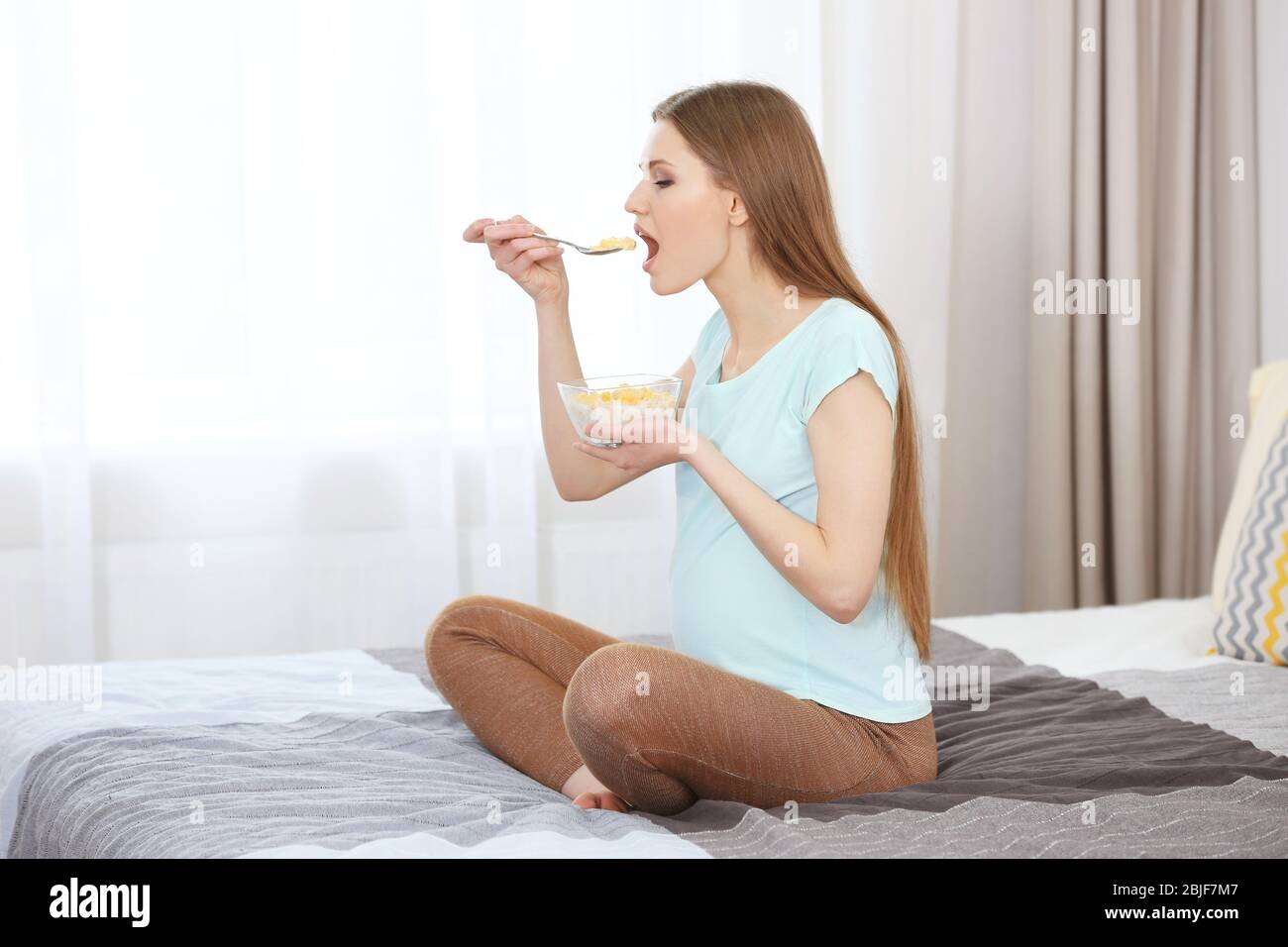 Beautiful pregnant woman eating cornflakes while sitting on bed at home