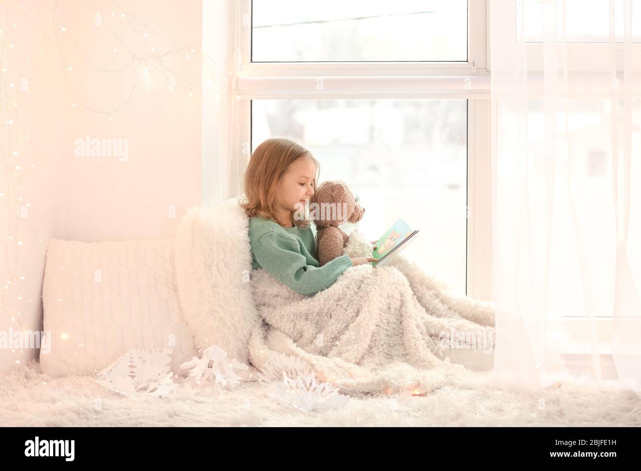 Cute little girl reading book while sitting on window sill at home ...