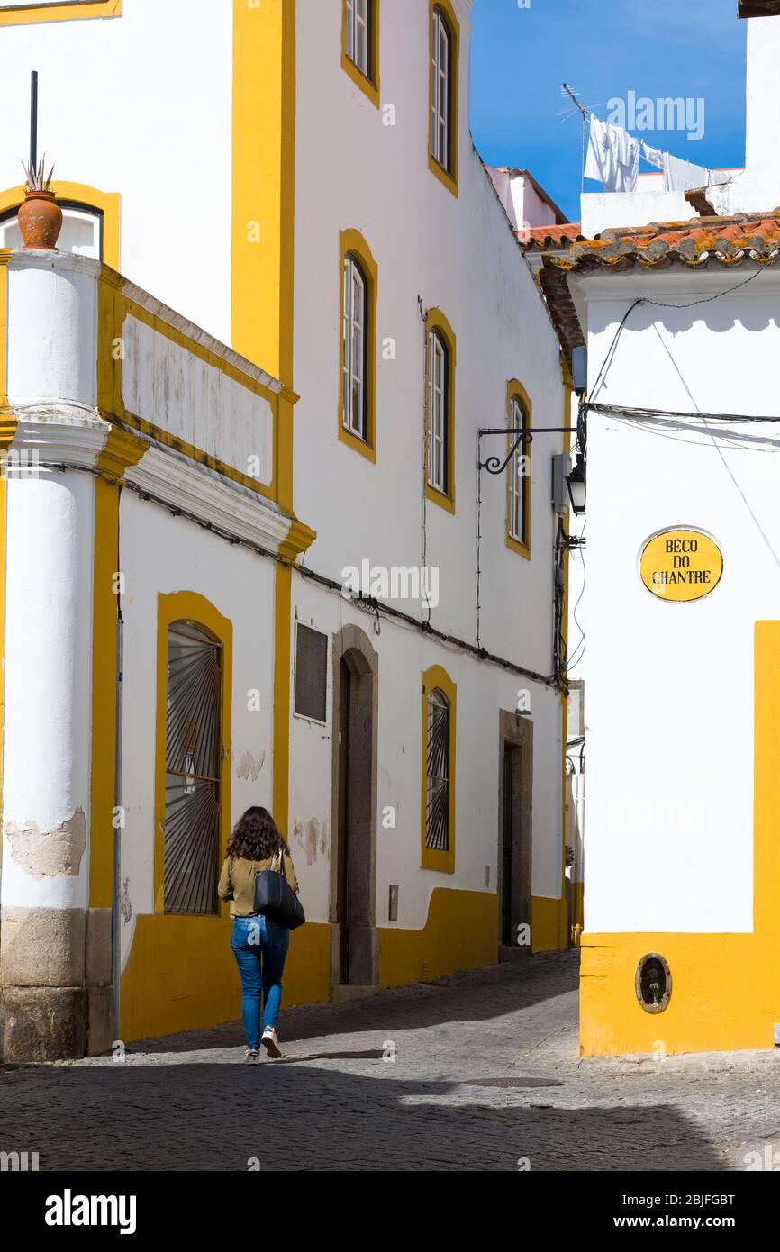 Stylish young woman walking in typical street scene of white and yellow houses, lanterns and narrow cobble street in Evora, Portu Stock Photo