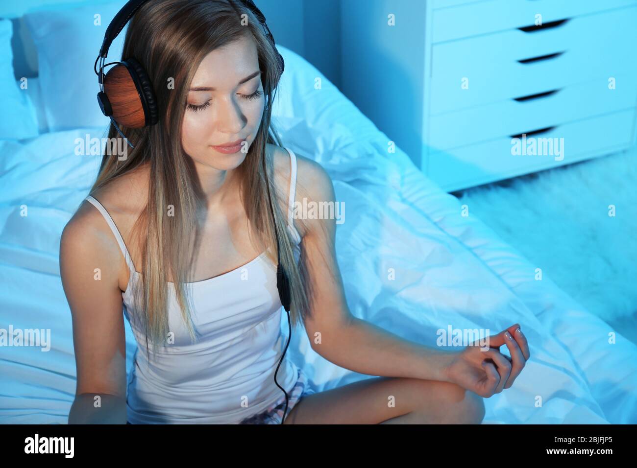 Beautiful young woman meditating and listening to music while sitting on bed at home Stock Photo