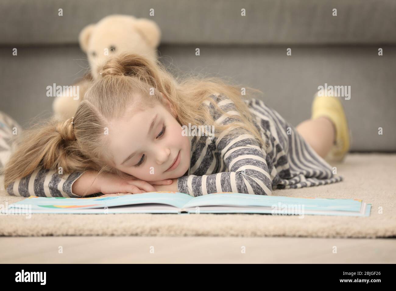 Small girl lying on floor near sofa and reading book in light room ...