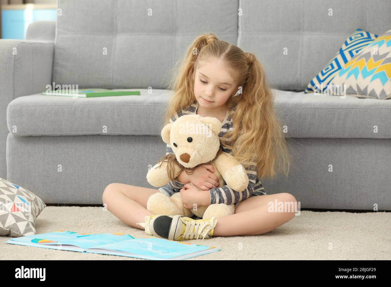 Small girl holding teddy bear and reading book near sofa Stock Photo ...