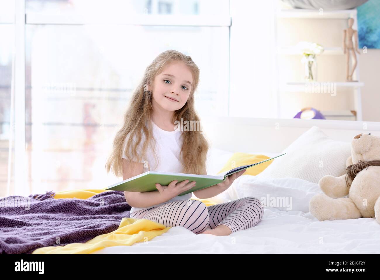 Small girl sitting on bed and holding book in light room Stock Photo ...
