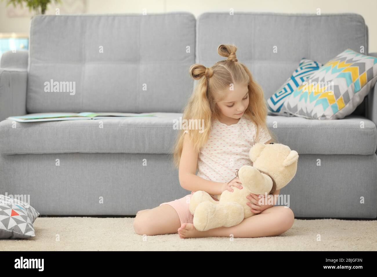 Small girl holding teddy bear and sitting near sofa in light room Stock ...